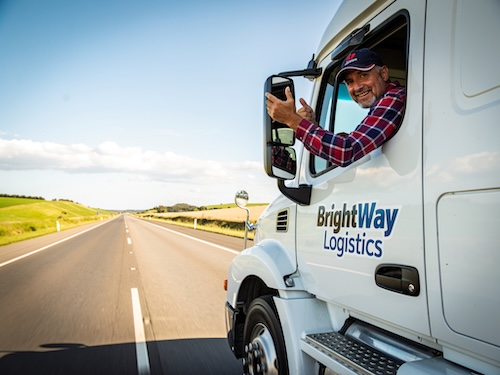 Fröhlicher LKW-Fahrer in einem sauberen, modernen LKW auf der Autobahn an einem sonnigen Tag, freut sich über seinen neuen Job.